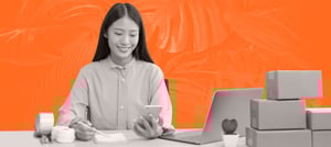 Mulher com um celular, em frente a um notebook e caixas de produtos, explorando a estratégia GTM para sua empresa - Tropical Hub